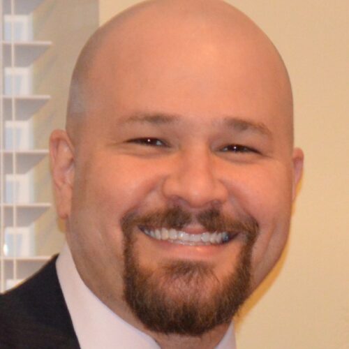 A smiling man with a bald head and a goatee, wearing a suit and tie, stands indoors near a window with blinds, discussing 2025 tech trends in Private 5G.