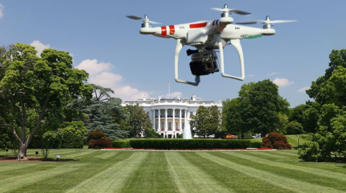 A drone equipped with Domestic Counter Unmanned Aircraft Systems hovers over the lawn in front of the White House on a clear day, with trees and a fountain visible in the background.