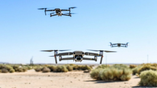 Three drones gracefully navigate a sandy desert landscape, their presence a testament to cutting-edge Domestic Counter Unmanned Aircraft Systems technology, all under the expansive stretch of a clear blue sky with sparse vegetation below.