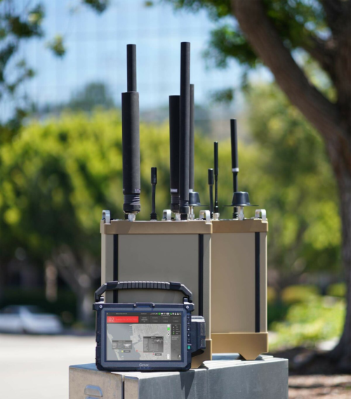 A Domestic Counter Unmanned Aircraft System sits on a concrete surface outdoors, featuring multiple antennas and a control device. Blurred trees and buildings create a dynamic backdrop for this sophisticated technology equipment.