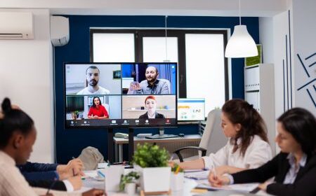 Four people sit at a conference table in an office equipped with advanced AV Solutions, participating in a virtual meeting with four others displayed on a large monitor screen.