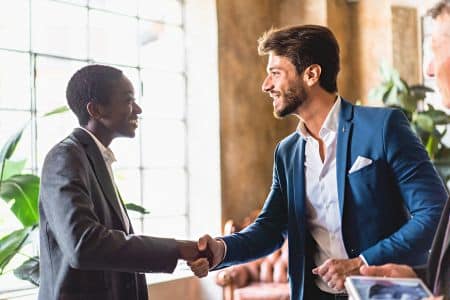 Two men in business attire shake hands and smile at each other indoors, with sunlight streaming through large windows and plants in the background—discussing innovative AV solutions for their workplace.