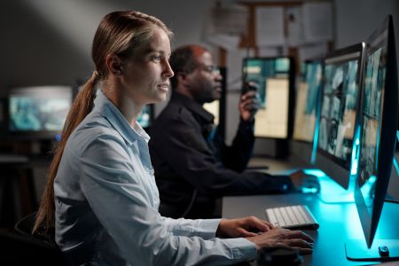 Two people sit at desks working on desktop computers with multiple monitors in a dimly lit office, utilizing advanced AV solutions for efficient workflow.
