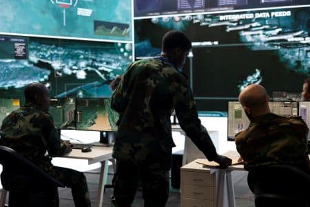 Three military personnel in camouflage uniforms analyze data and maps on multiple computer screens, utilizing advanced AV solutions in a command center with large digital displays.
