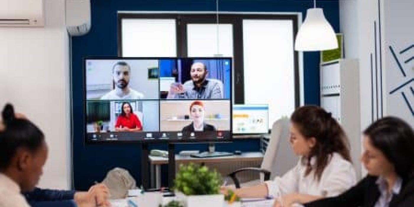 Four people sit at a conference table in an office equipped with advanced AV Solutions, participating in a virtual meeting with four others displayed on a large monitor screen.